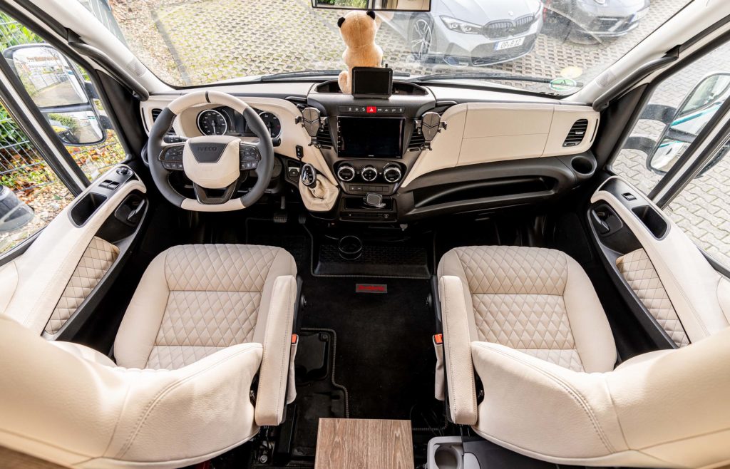 Das Fahrerhaus des Wohnmobils mit zwei bequemen Sitzen mit beigefarbenem, gestepptem Leder, einem Teddybären auf dem Armaturenbrett und modernen Fahrzeugsteuerungen.